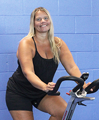 Female spin instructor in a black tank top poses on a spin bike in front of a blue wall