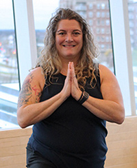 Female yoga instructor stands with hands in prayer pose in front of a window. She is wearing a black tank top